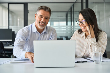 2 multicultural team coworkers, older Latin man executive and young Asian woman manager working together, happy diverse employees looking at laptop computer discussing financial corporate project.