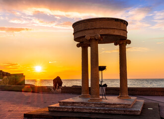 Fototapeta premium beautiful stone monument with three columns in ancient history greek style with see embarkment and amazing cloudy sunset on background of landscape
