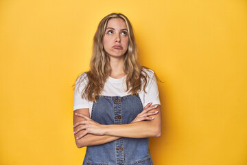 Young blonde Caucasian woman in denim overalls posing on a yellow background, tired of a repetitive...