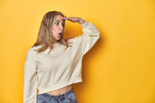 Young Blonde Caucasian Woman In A White Sweatshirt On A Yellow Studio Background, Looking Far Away Keeping Hand On Forehead.