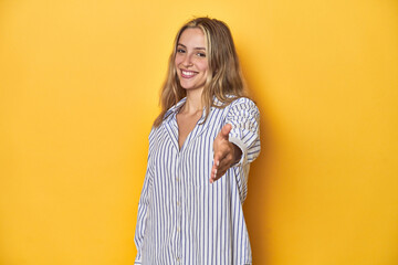 Young blonde Caucasian woman in a striped business shirt on a yellow background, stretching hand at...