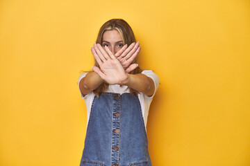 Young blonde Caucasian woman in denim overalls posing on a yellow background, doing a denial gesture