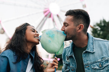 Young guys are having fun in an amusement park. Girlfriend and her boyfriend biting cotton candy...