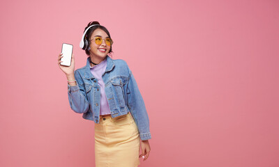 Happy smile Asian teen woman showing blank screen smartphone she listening music in headphones isolated on pink copy space background.