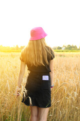 Back view of pretty tall girl standing in the yellow wheat field. Sunny day. Pink panama hat. Vertical. Purple phone in the pocket