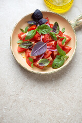 Sliced red tomatoes with fresh basil and olive oil in a serving pan, vertical shot on a beige stone background with space, elevated view
