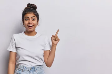 Tuinposter Horizontal shot of pretty surprised cheerful young woman pointing to empty copy space advertises product or tells about awesome offer dressed in casual clothing isolated over white background © Wayhome Studio