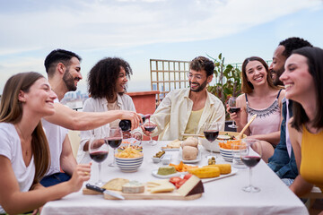 Group of multiracial friends gathering in a rooftop during daytime, eating and drinking enjoying the good weather. People having fun in a terrace, drinking wine and sitting in a table full of food. 