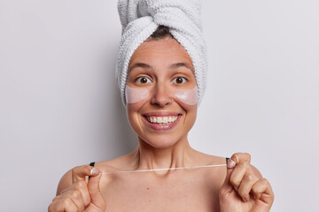Positive European woman smiles broadly poses with towel wrapped on head holds dental floss going to...