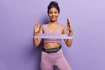 Athletic woman in sportswear showcases strength as she stretches rubber resistance band her radiant smile reflects commitment to fitness and healthy lifestyle isolated over purple background.