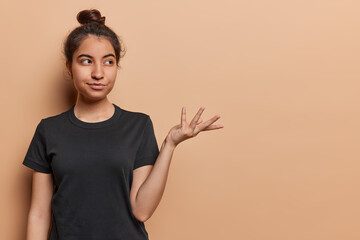 Horizontal shot of beautiful Iraniam girl with dark hair gathered in bun keeps palm raised up over blank space against brown background wears casual black t shirt attracts your attention to something