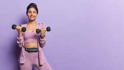 Motivated sporty woman showcases her strength as lifts dumbbells engaging in challenging workout dressed in sportswear isolated over purple background copy space for your promotional content