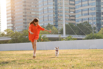 Teenager girl playing with small white dog Jack Russell terrier. adorable friends having fun running in the field. Summer in big city