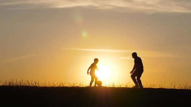 Silhouette of a man and a child at sunset. A boy is playing soccer with his dad. Male coach. Games with children, active recreation. The concept of a child dream