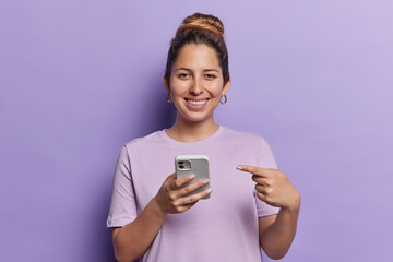 Fototapeta premium Horizontal shot of pretty young female model holding cellphone advertising new trendy application points at modern gadget smiles toothily wears casual t shirt isolated over purple background