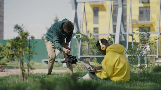 Young african guy is filmed in video on territory of residential complex. Cameraman shoots guy in yellow hoodie with headphones and laptop on lawn in yard of house. Slow motion. Backstage