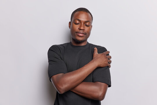 Dark skinned man wearing casual black t shirt envelops himself in loving embrace his eyes closed exudes sense of contentment and gratitude finding comfort in his own presence isolated over white wall