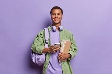 Smiling African teenage student dressed in trendy attire confidently holds books while engrossed in mobile phone conversation showcasing modern integration of technology in education purple background