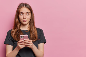 Horizontal shot of pretty young woman indulges in her daydreams engrossed in thoughts holds mobile...