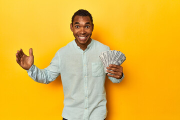 African American man with dollar bills, yellow studio, receiving a pleasant surprise, excited and...