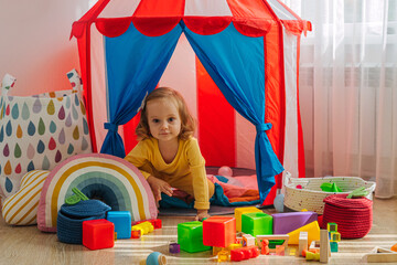 A little girl sitting on floor plays with colorful toys in playroom. Educational game for baby and...