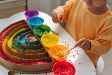 Child hands playing colored rice and make rainbow.  Child filled the rainbow with bright rice. Montessori material. Sensory play and learning colors activity for kids.