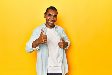 African American man in blue shirt, yellow studio, raising both thumbs up, smiling and confident.