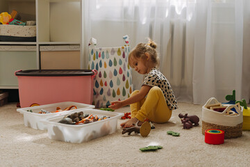 A little girl playing with toy animals and wooden blocks in nursery. Kid and toys with storage...