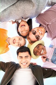Vertical of Multiracial and divers group of friends hugging in circle and looking down at the camera. They stand happy outside. Putting their heads together. Low angle of a young people taking selfie