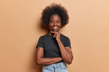Fototapeta na wymiar Waist up shot of dark skinned woman touches chin gently smiles broadly dressed in casual black t shirt and jeans expresses positive emotions isolated over brown wall. Satisfied African female model