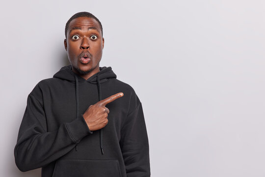 African man stands with look of utter astonishment pointing towards right copy space demonstrates advertisement dressed in casual black hoodie isolated over white background. Omg look there.