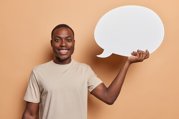 Positive friendly dark skinned man holds empty speech bubble brimming with anticipation to convey his heartfelt thoughts dressed in casual t shirt isolated over brown background. Place your promo here