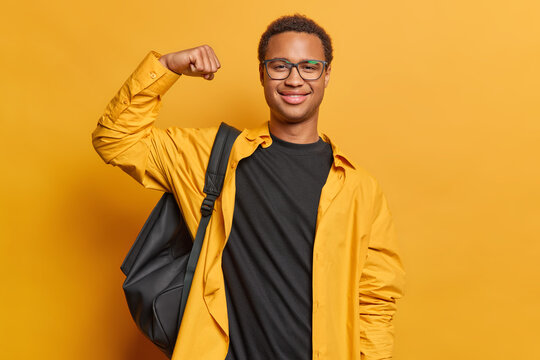 Cheerful African male student brags about his personal achievements raises arm shows biceps carries rucksack wears shirt and black t shirt isolated over yellow background. Look how strong I am