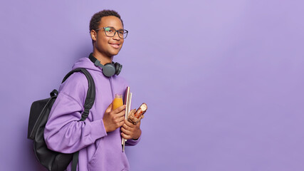 Back to school. Sideways shot of pleased dark skinned guy poses with notepads and food for snack enjoys studying carries rucksack poses against purple background with copy space for your advertisement