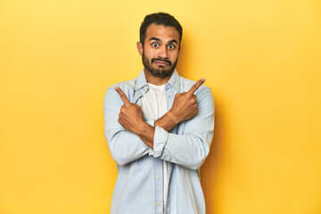 Casual young Latino man against a vibrant yellow studio background, points sideways, is trying to choose between two options. - obrazy, fototapety, plakaty