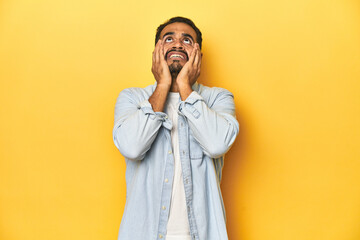 Casual young Latino man against a vibrant yellow studio background, whining and crying disconsolately.