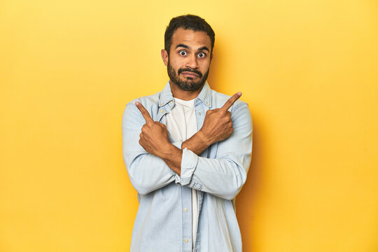 Casual young Latino man against a vibrant yellow studio background, points sideways, is trying to choose between two options.