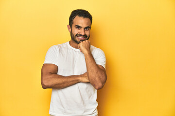 Casual young Latino man against a vibrant yellow studio background, smiling happy and confident, touching chin with hand.