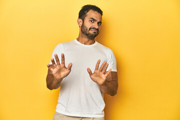 Casual young Latino man against a vibrant yellow studio background, rejecting someone showing a...