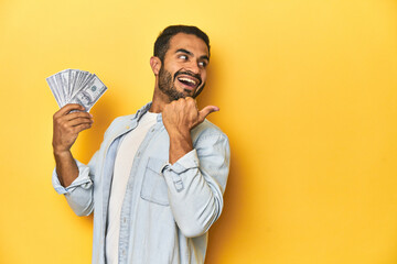 Young Latino man holding a bundle of dollars, yellow studio background, points with thumb finger...