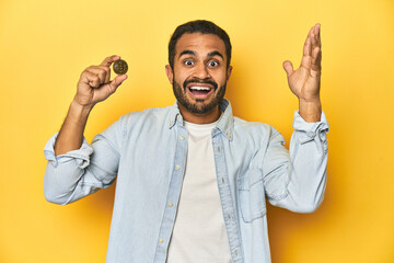Young Latino man holding a Bitcoin coin, yellow studio background, receiving a pleasant surprise,...