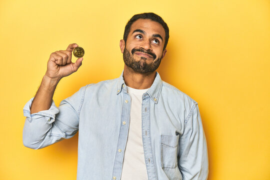Young Latino man holding a Bitcoin coin, yellow studio background, dreaming of achieving goals and purposes