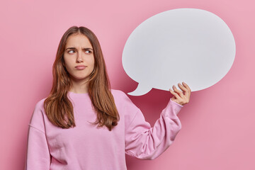 Horizontal shot of displeased young European woman with long straight hair frowns face dressed in casual pullover holds communication bubble for your advertisement isolated over pink background.