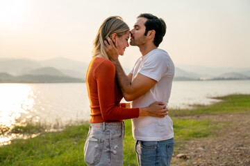 Caucasian man kiss on forehead of beautiful woman with sunset light near lake and look romantic for couple love stay together.