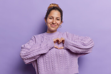 I like it. Tender smiling pretty woman showing heart gesture has happy face expression dressed in casual knitted sweater isolated over purple background adores something likes product or person
