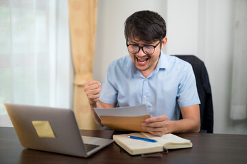 A handsome young man in glasses succeeds while reading acceptance letter with a laptop on his desk at home.