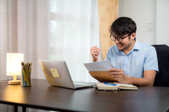 A Handsome Young Man In Glasses Showing Success Gesture While Reading Acceptance Letter With Laptop On Desk At Home