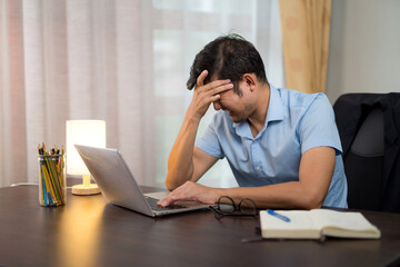 Asian man having severe migraine headache due to long hours of work and reserving a laptop at his desk at home.