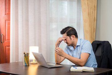 Stressed Asian man taking off his glasses and pinching his eyebrows on his home desk with a laptop.