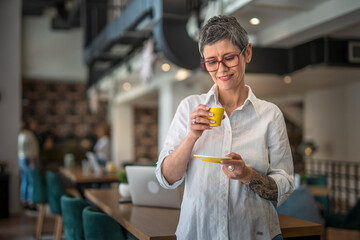 One senior woman with tattoo on left hand standing at cafe and drinking coffee from yellow cup,...